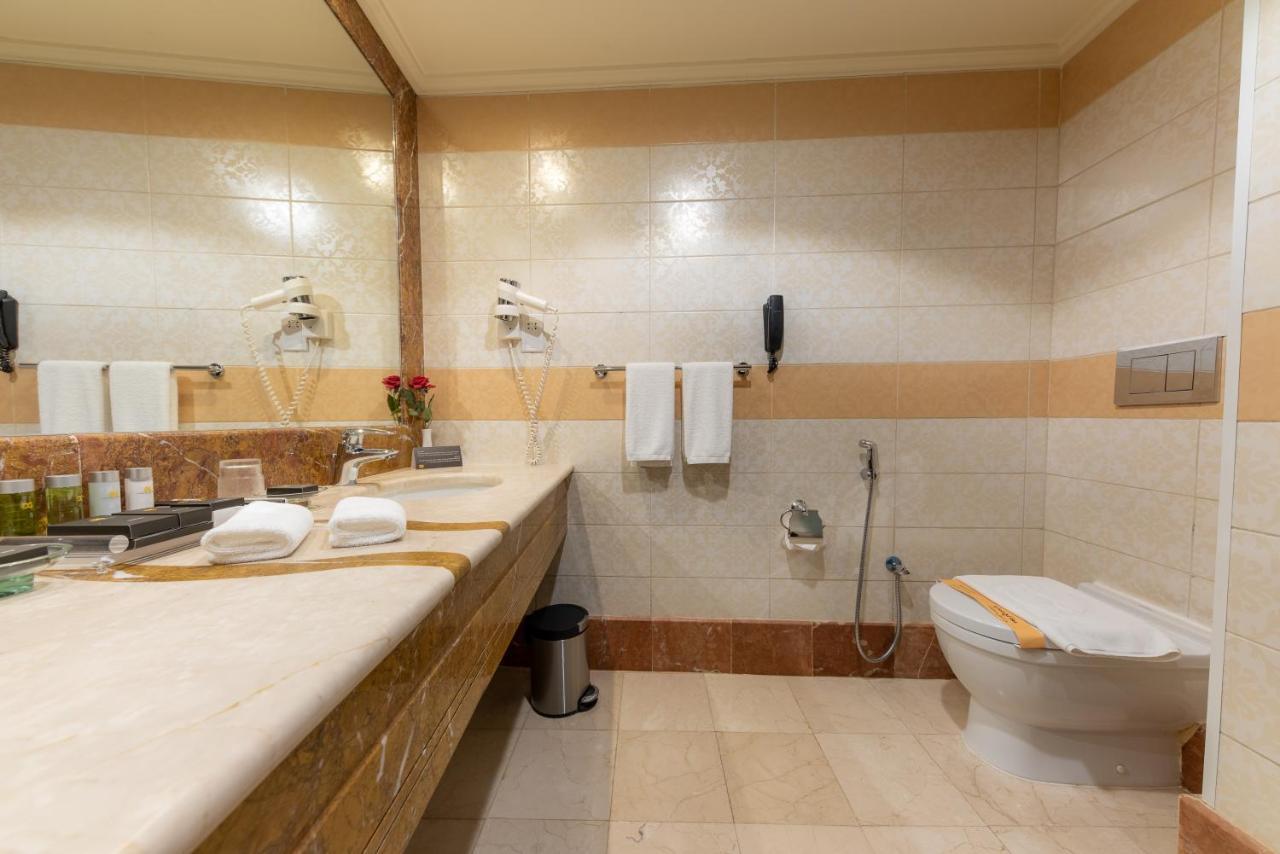 Al Ghufran Safwah Hotel Makkah La Meca Exterior foto The photo shows a bathroom that features a large countertop with a sink, which is made of a light-colored stone. There are several toiletries and a small vase with flowers on the countertop. Above the sink, there's a large mirror reflecting the space