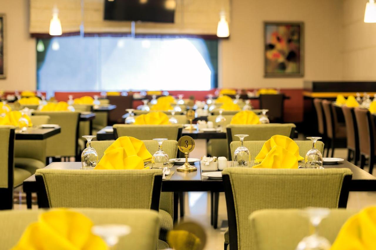Al Ghufran Safwah Hotel Makkah La Meca Exterior foto The photo shows a restaurant interior with several dining tables arranged in a spacious setting. Each table is set with glasses, utensils, and folded yellow napkins. The chairs have a green upholstery, and there's ambient lighting with hanging fixtur